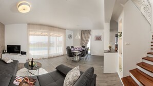 Una sala de estar moderna con un sofá, una mesa de centro y un televisor. Se observa un comedor con una mesa y sillas. Se ve una escalera con escalones de madera.