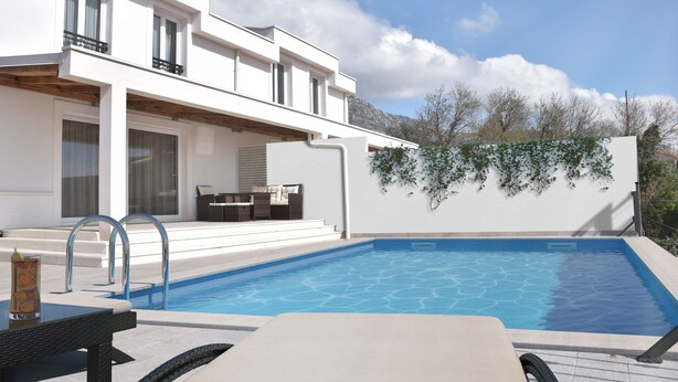 Un área moderna junto a la piscina con una piscina azul cristalina, un edificio blanco con balcones y montañas al fondo.
