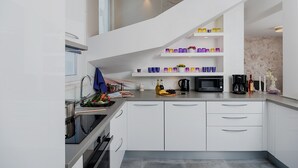 Una cocina moderna con armarios blancos, un lavamanos de acero inoxidable y un microondas integrado. Hay estantes sobre la encimera con tazas de colores y una cafetera.