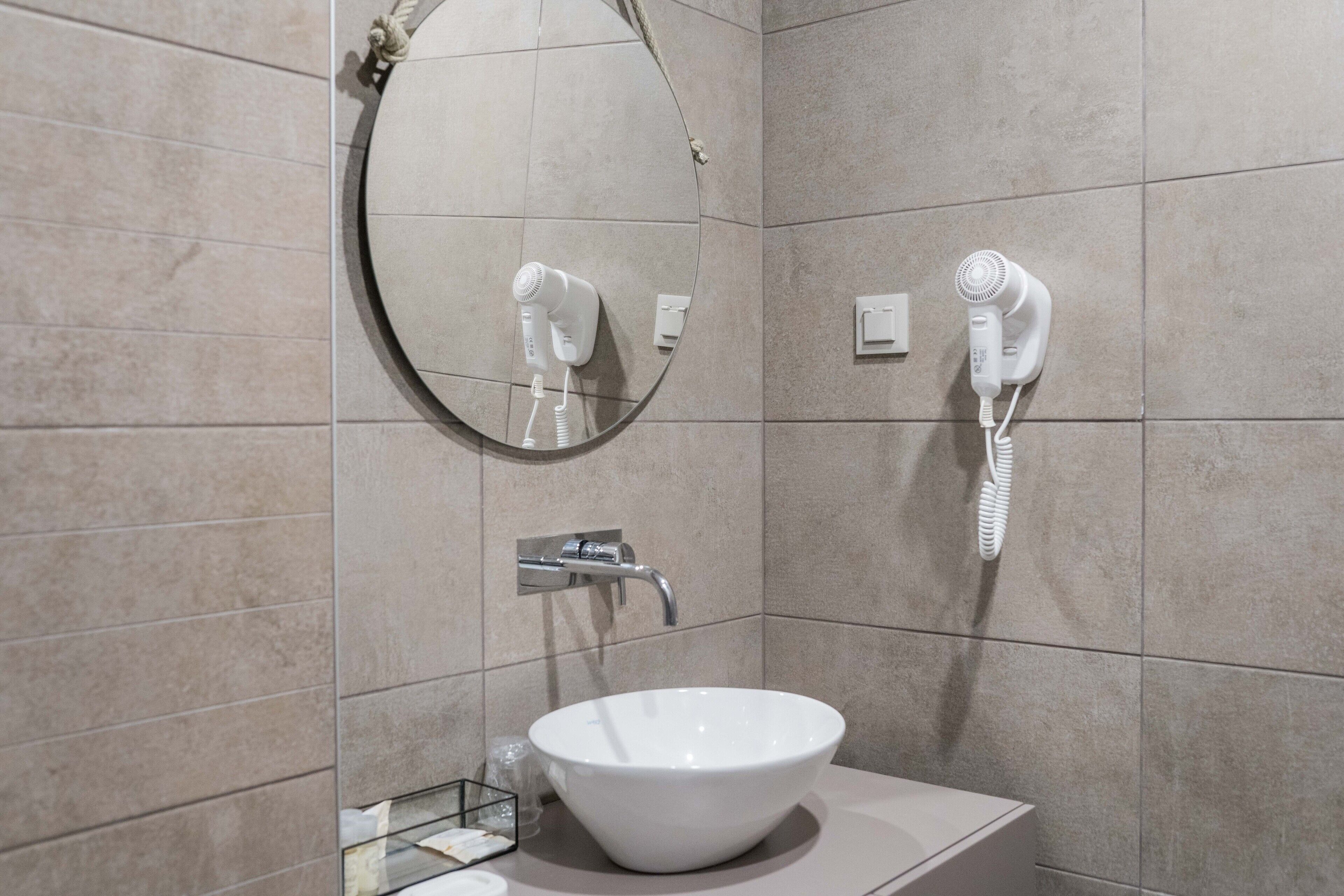 Une salle de bain moderne avec un miroir rond, un lavabo blanc et deux sèche-cheveux fixés au mur.