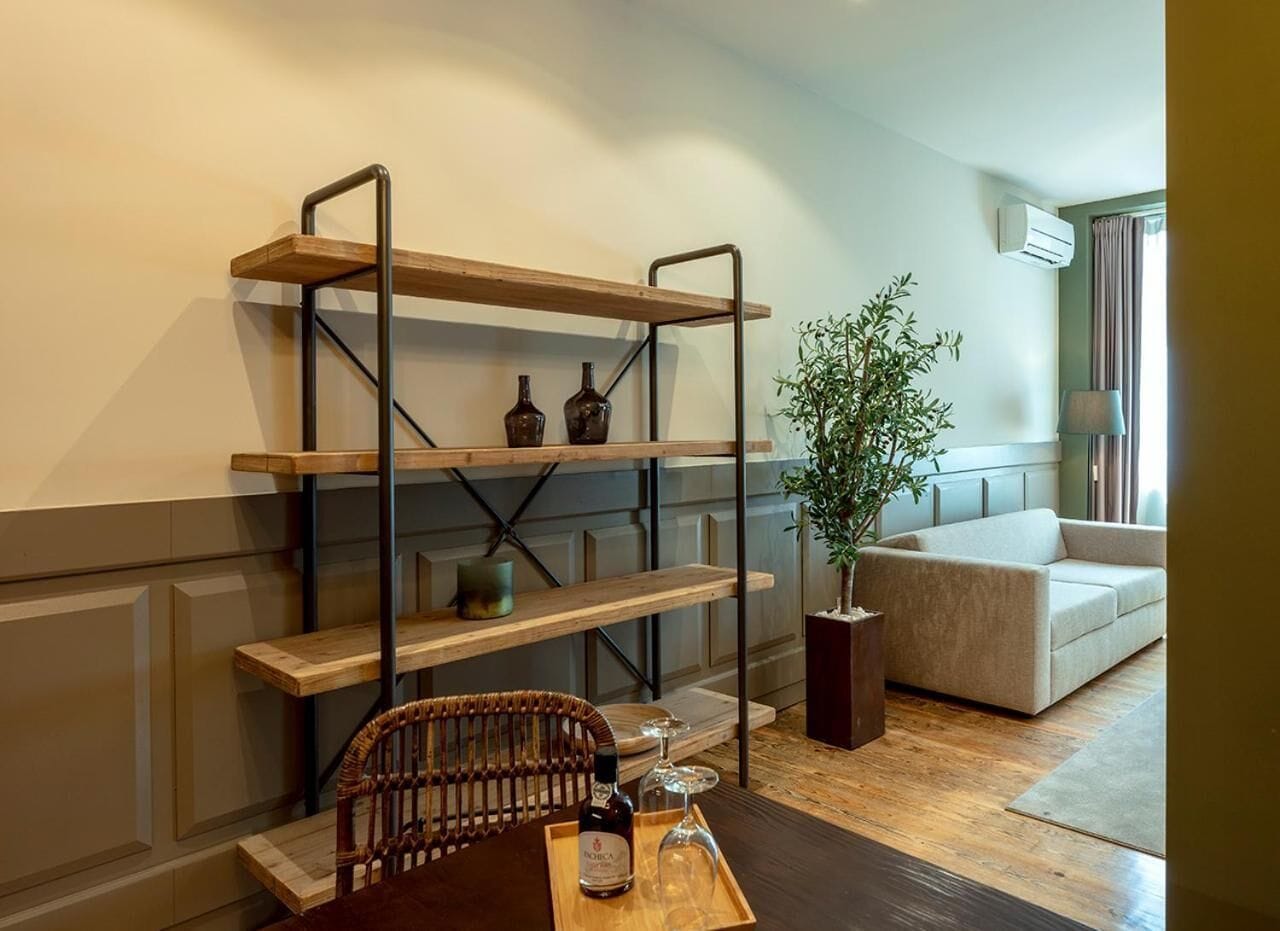 A living room with a wooden coffee table, wicker chair, and a built-in wooden shelf unit with bottles and a plant.