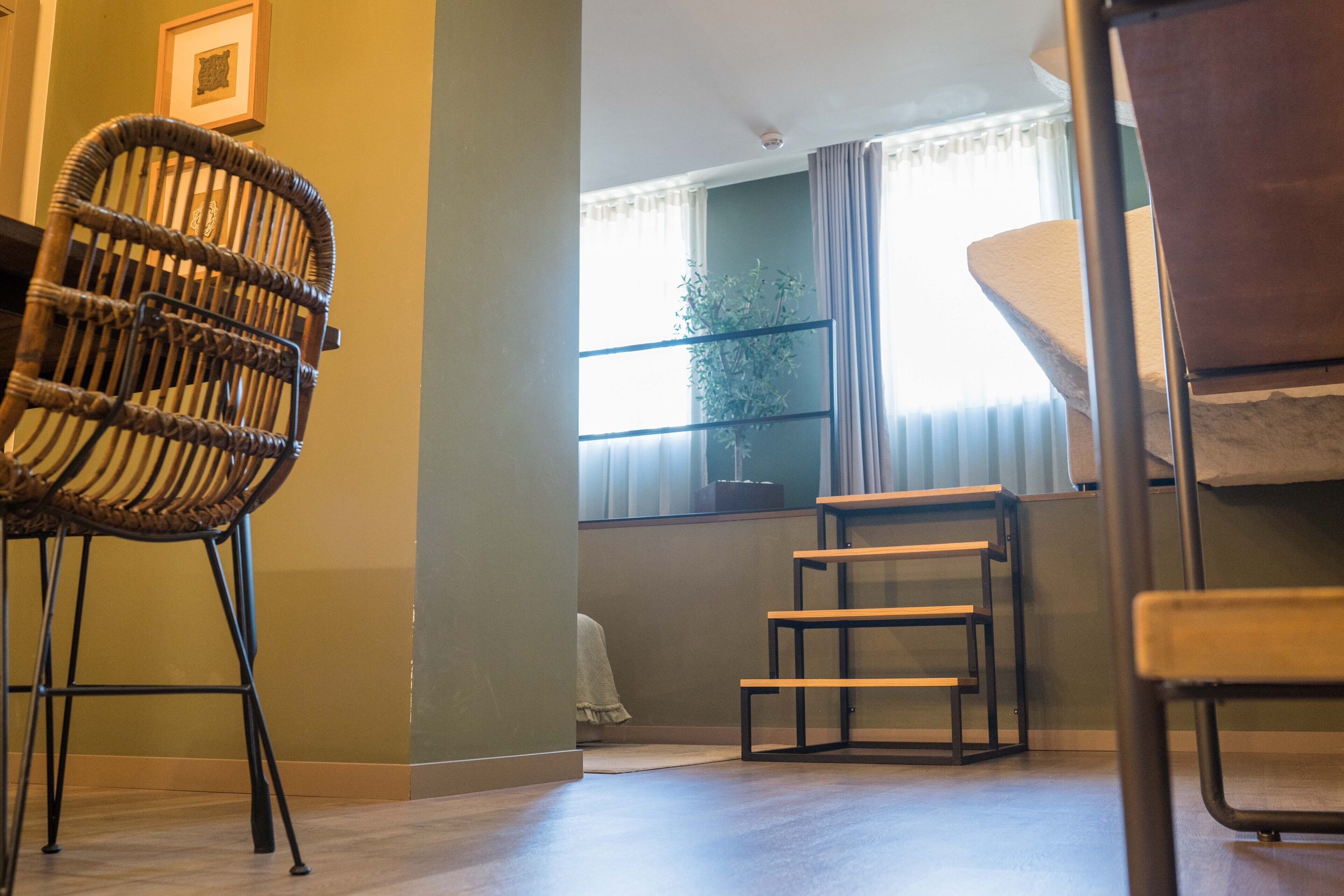 Ein Zimmer mit einem Korbsessel, einem Holzhocker und einem Bett mit weißer Bettdecke.