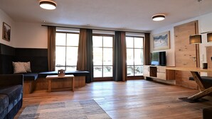 A modern living room with a sofa, coffee table, and TV. There are large windows with curtains, a wooden cabinet, and a rug on the floor. - Residence Martha (Biberwier)