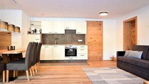 A modern kitchen with white cabinets, a built-in oven, and a wooden accent wall. There is a dining area with a wooden table and chairs, and a gray rug in the living room with a dark sofa. - Residence Martha (Biberwier)