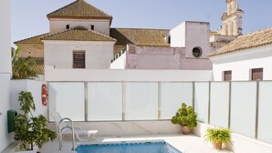 Une piscine rectangulaire à l’eau d’un bleu limpide, entourée de murs blancs et surmontée d’un toit en tuiles. Des chaises longues blanches et des plantes en pot sont disposées autour de la piscine.