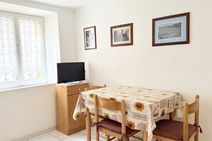 Un área de comedor con una mesa cubierta por un mantel estampado, sillas de madera, un televisor sobre un mueble de madera y tres cuadros en la pared.