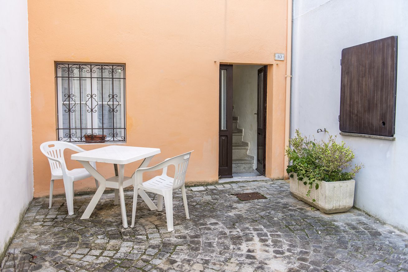 A narrow alleyway with a white table and chairs, a potted plant, and a door leading to a building.