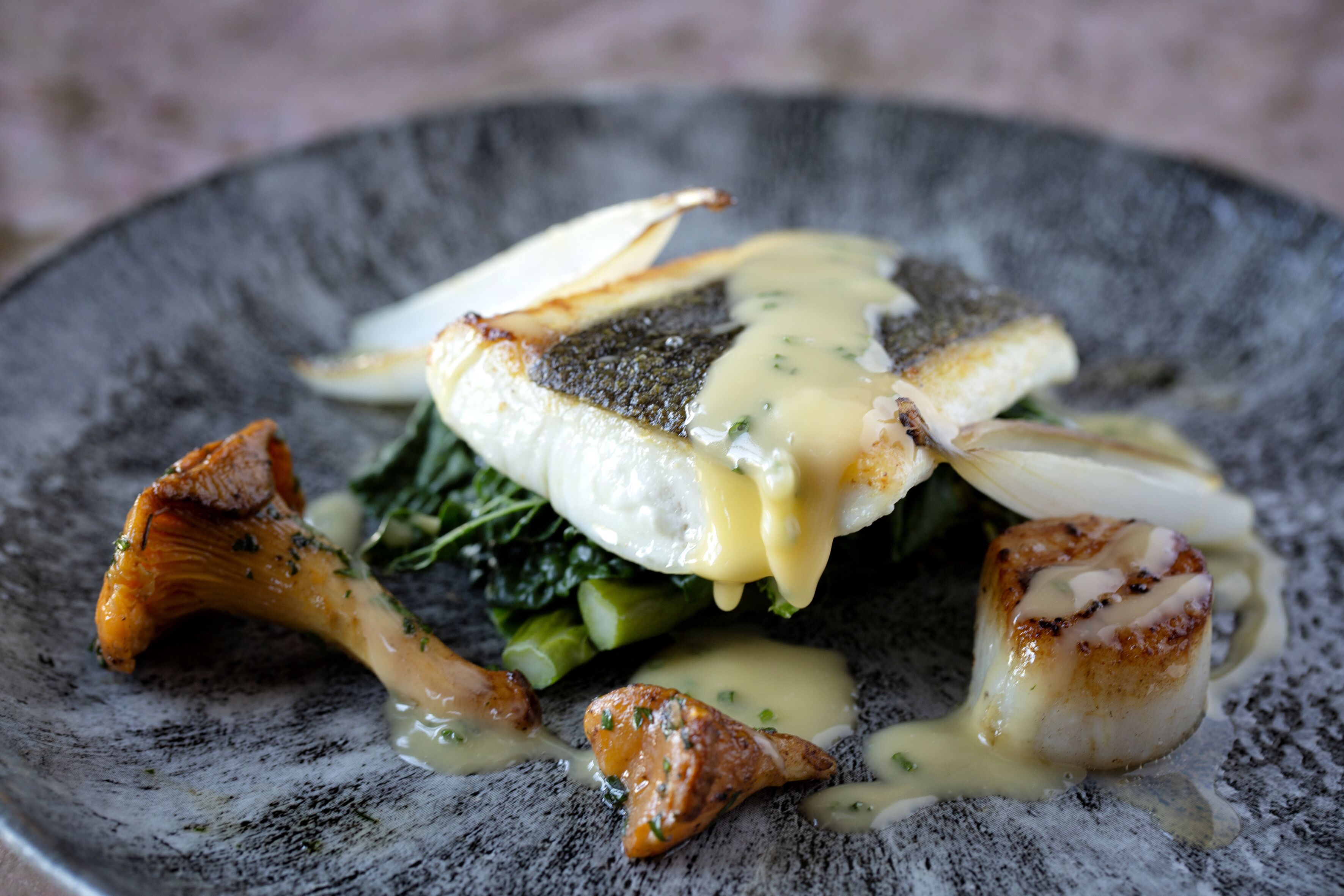 A plate of food with seared fish, mushrooms, and greens, served on a textured gray plate.