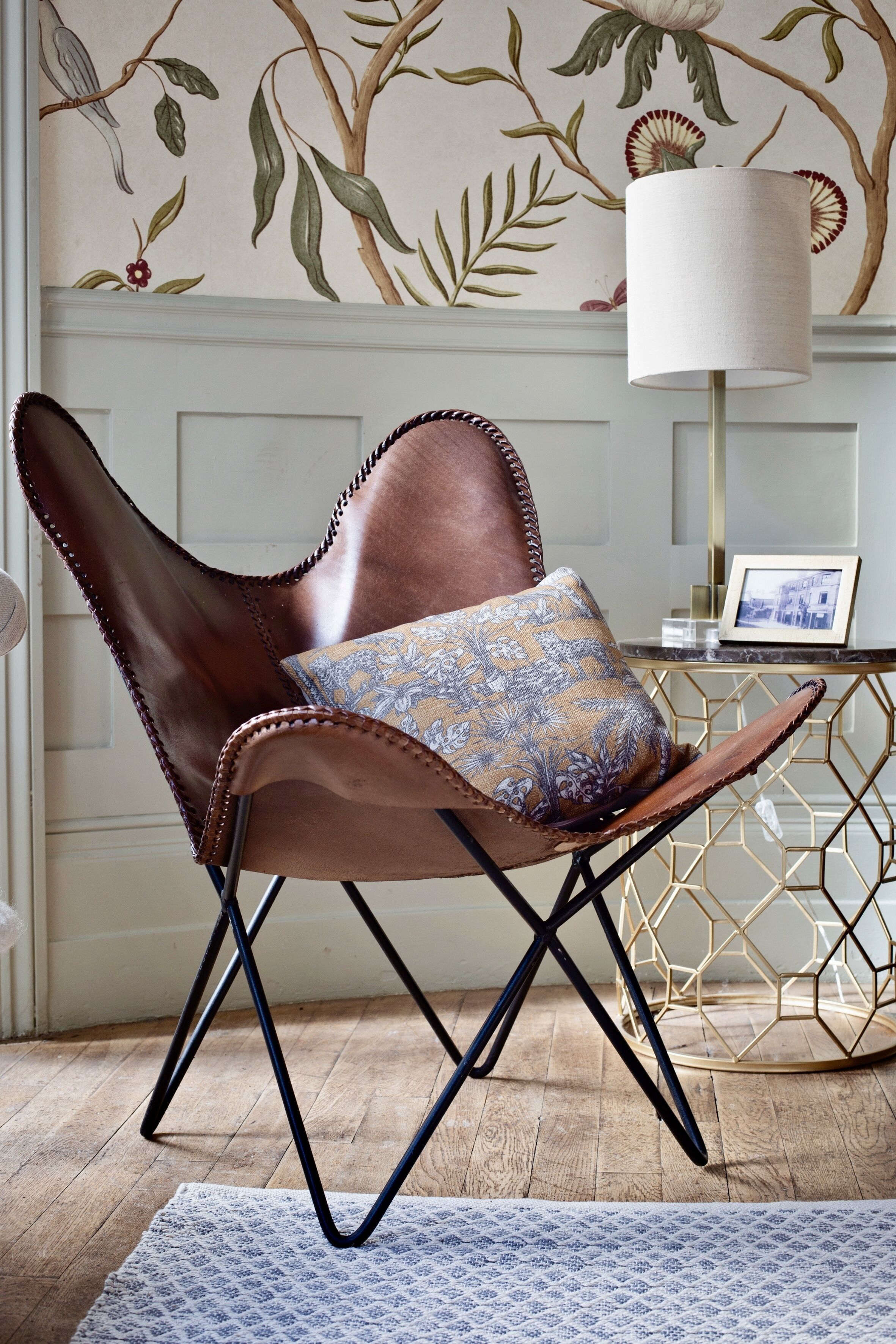 A leather chair with a patterned cushion, a gold geometric side table, and a wall with floral wallpaper.