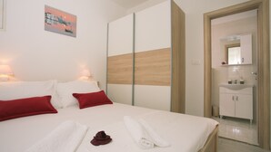 A hotel room with a bed, red pillows, white towels, a wooden accent wall, a painting, and a bathroom with a sink and mirror. - Apartments Kampanel (Kastela)