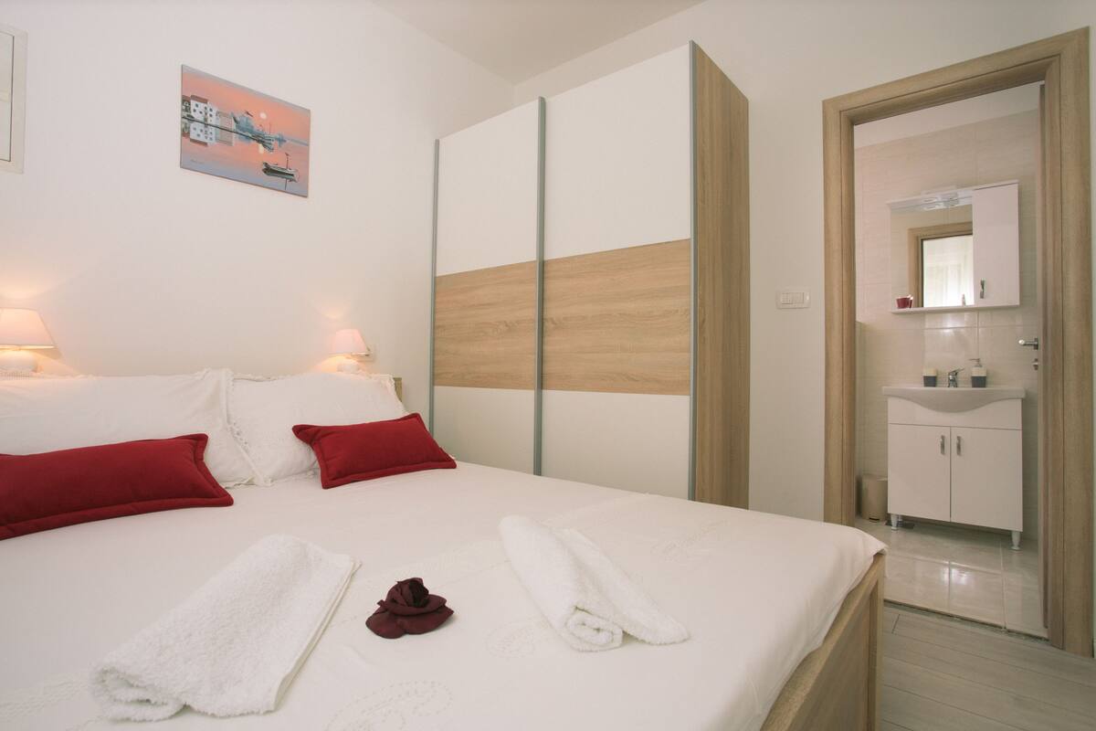 A hotel room with a bed, red pillows, white towels, a wooden accent wall, a painting, and a bathroom with a sink and mirror.