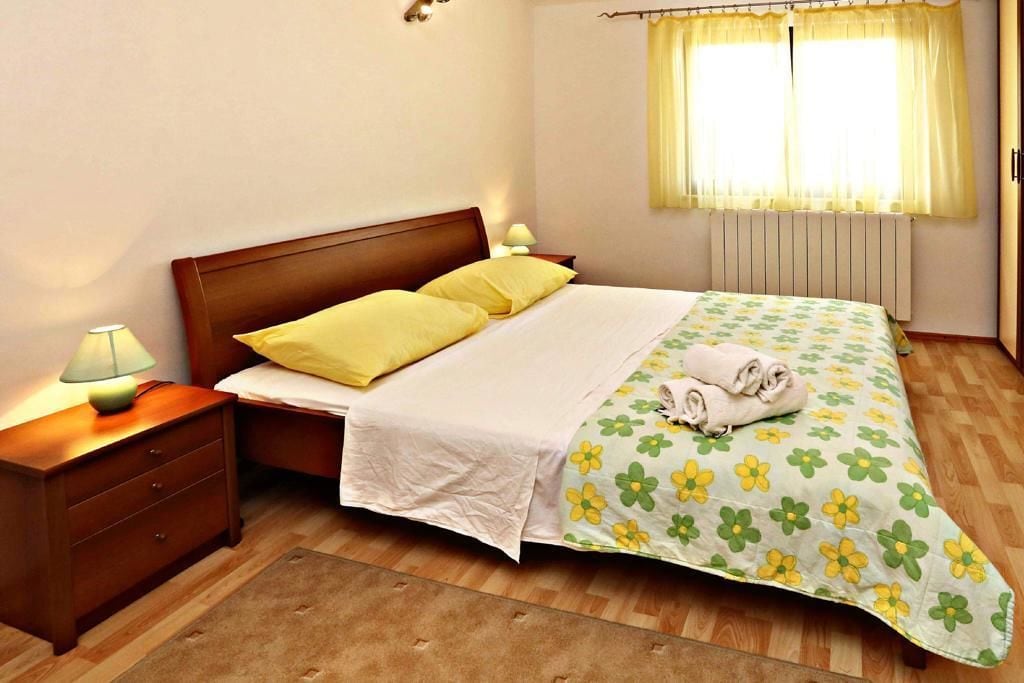 Ein ordentlich bezogenes Bett mit floralem Überwurf, ein Nachttisch aus Holz mit einer Lampe und ein Fenster mit Vorhängen.