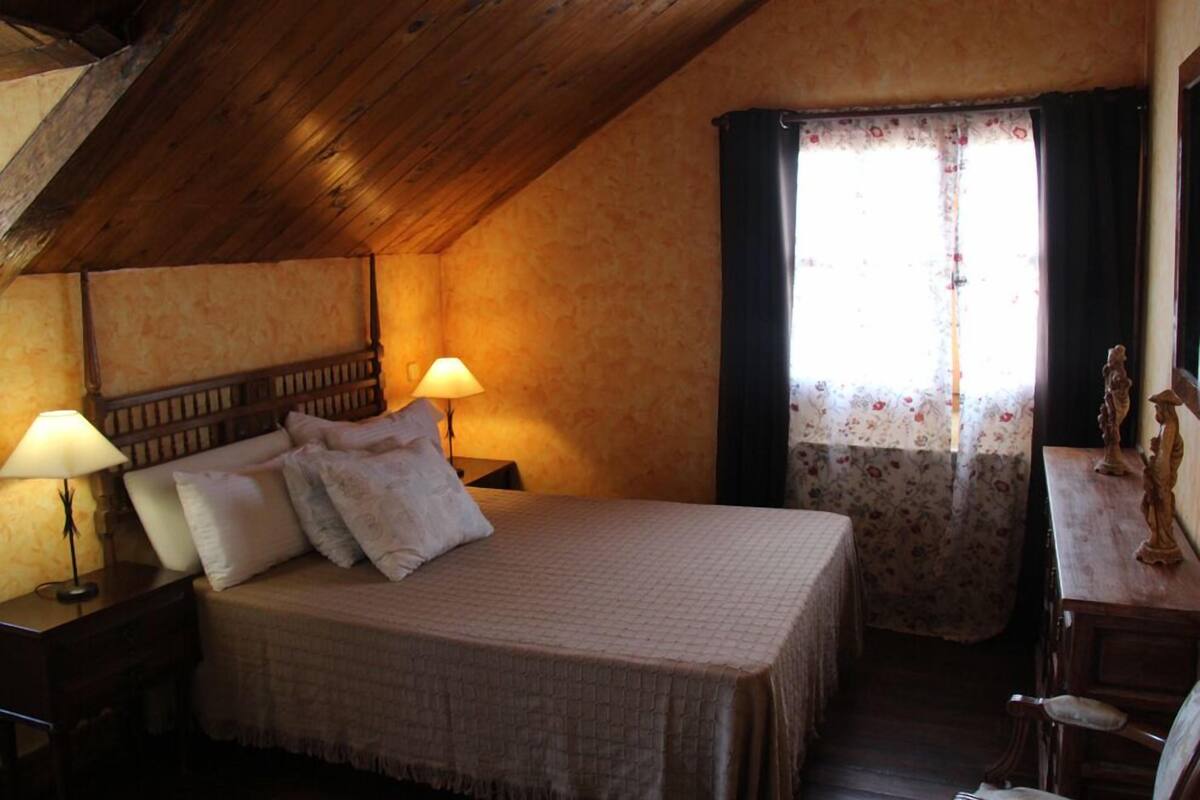 Un dormitorio acogedor con techo de madera, una cama con manta acolchada, dos lámparas de noche y una ventana con cortinas florales.
