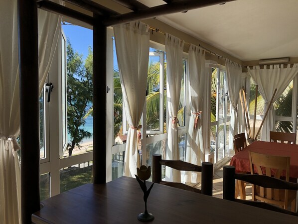 A room with large windows, a wooden table, and a view of a beach and palm trees. - Residence VMJ villa 6 @ (Trou D’eau Douce)