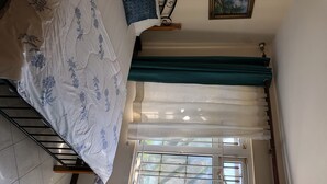 Ein Schlafzimmer mit einem Bett, einem Fenster mit Vorhängen, einem Heizkörper und Blick auf Grünfläche.
