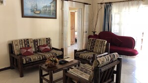 Ein Wohnzimmer mit zwei Sofas, einem Couchtisch und einem Sessel. An der Wand hängt ein Gemälde, und ein Fenster mit Vorhängen ist zu sehen.