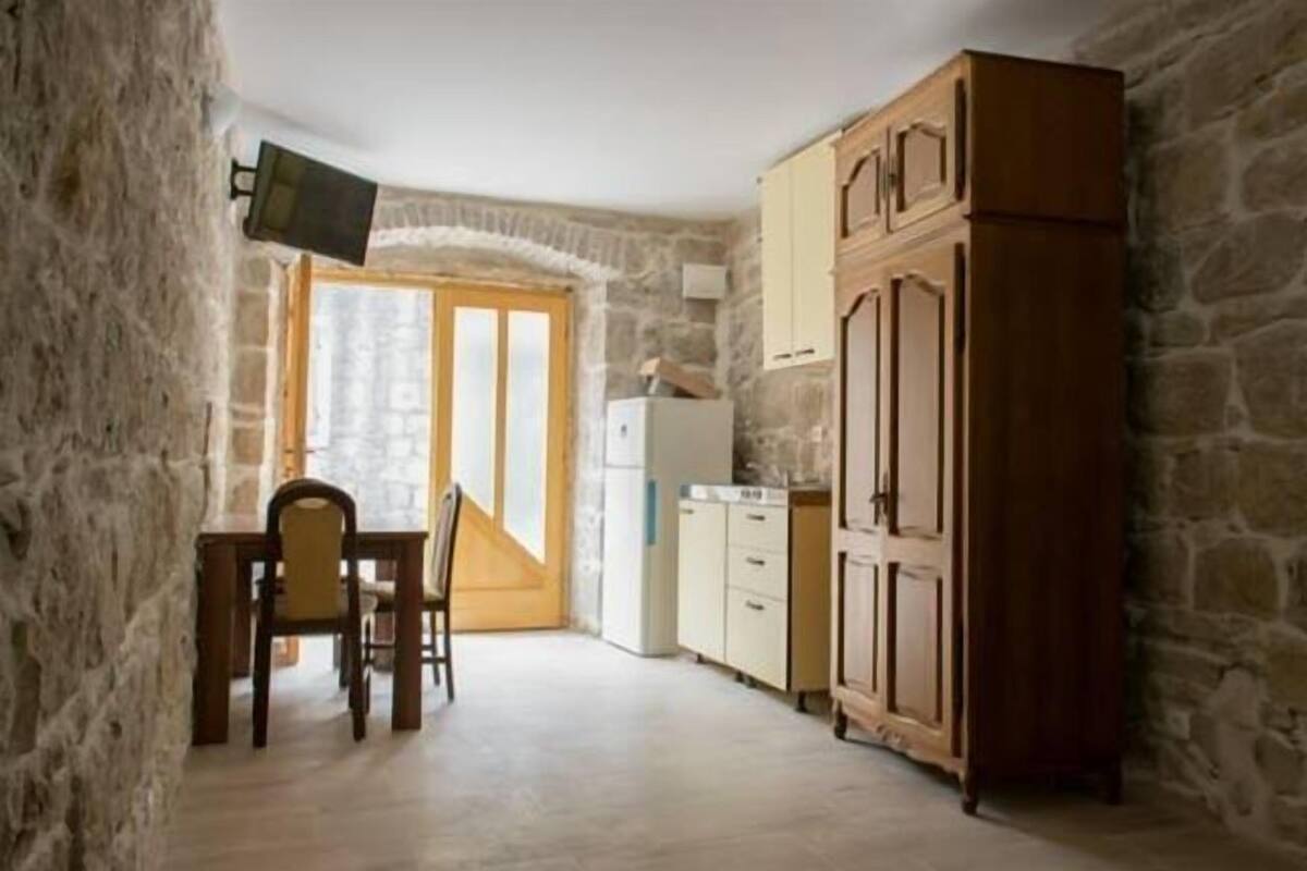A small kitchen with a stone wall, wooden cabinets, a dining table with chairs, and a television mounted on the wall.