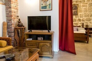 A cozy living room with a stone wall, a flat-screen TV, a wooden cabinet, a bed with a red curtain, and a painting on the wall.