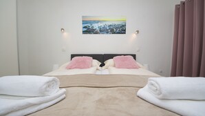 A neatly made bed with a beige blanket and two pink pillows, a painting of a beach scene on the wall, and a window with curtains.