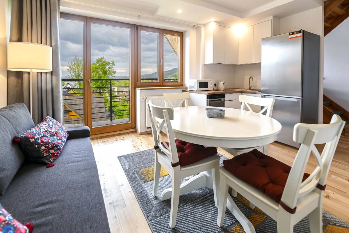 Una cocina moderna con armarios blancos, un refrigerador de acero inoxidable y una zona de comedor con sillas blancas y una mesa redonda.