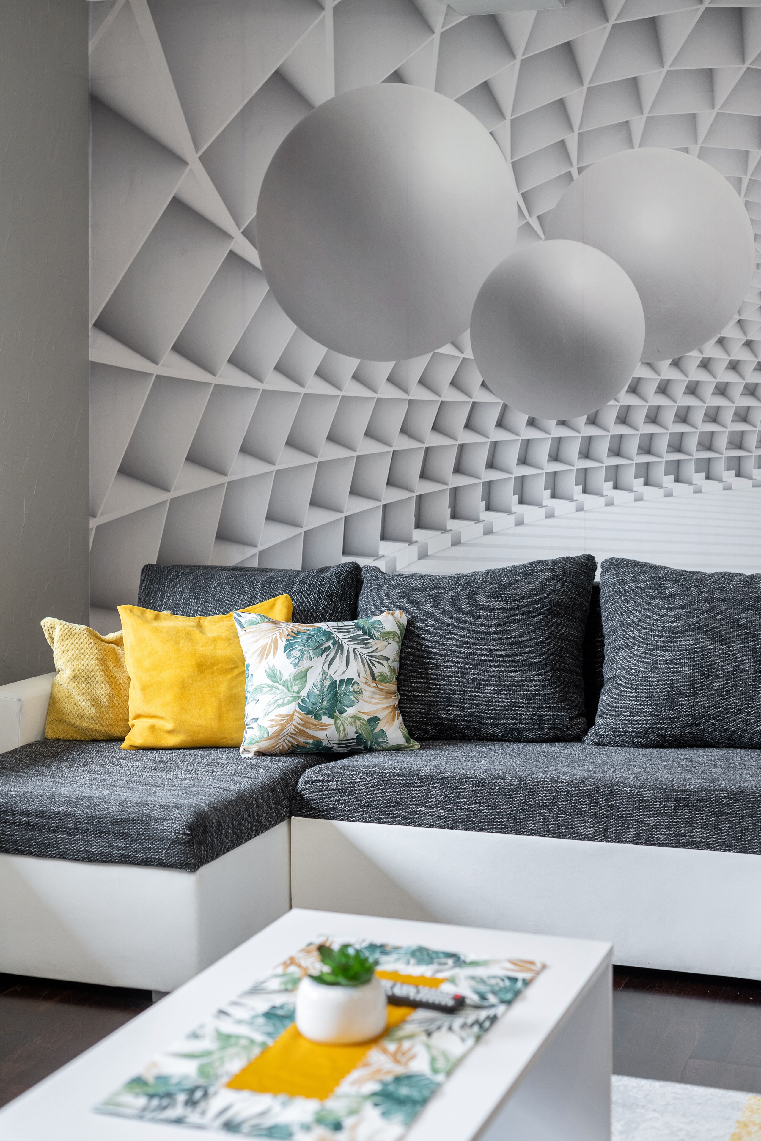 A modern living room with a geometric patterned wall, a sectional sofa with multiple cushions, and a coffee table with a plant.