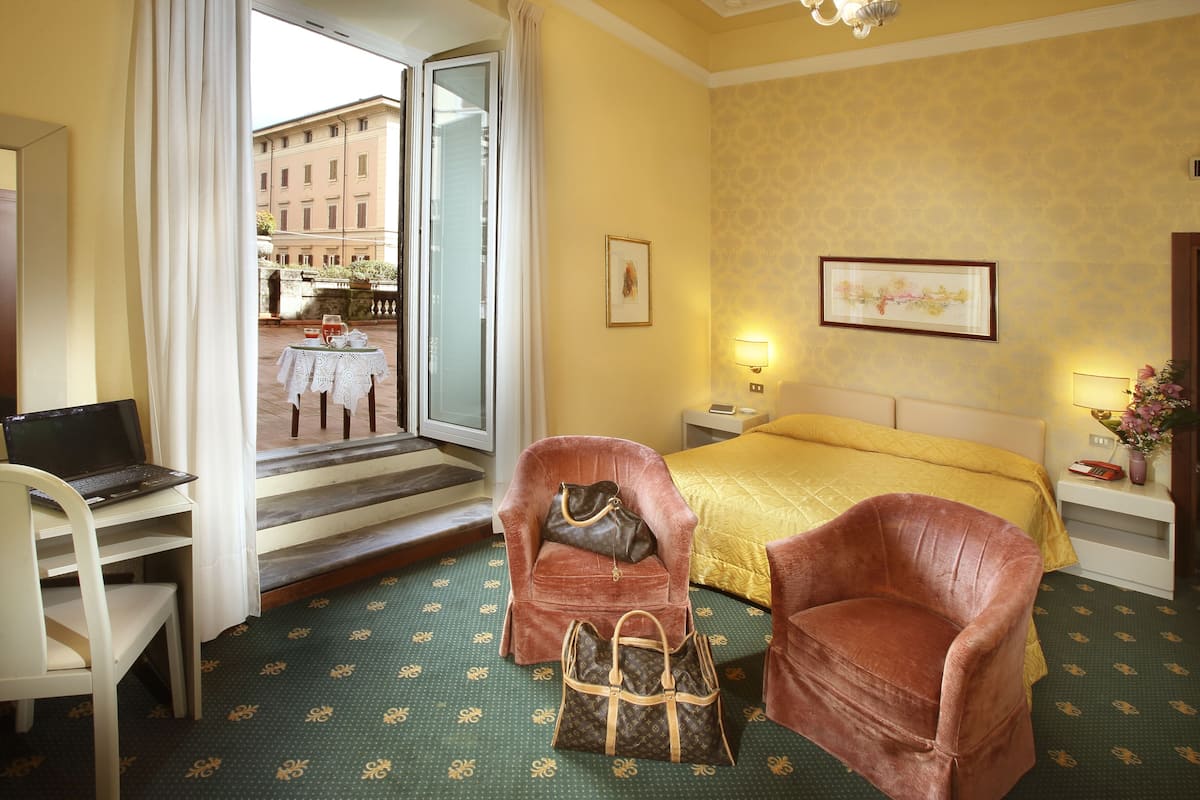 Habitación de hotel con cama, dos sillones, un escritorio con una computadora portátil, una ventana con cortinas, una mesita de noche con lámpara y vista a la calle.
