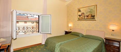 A hotel room with a bed, a nightstand, a lamp, a painting, and a window with a view of buildings.
