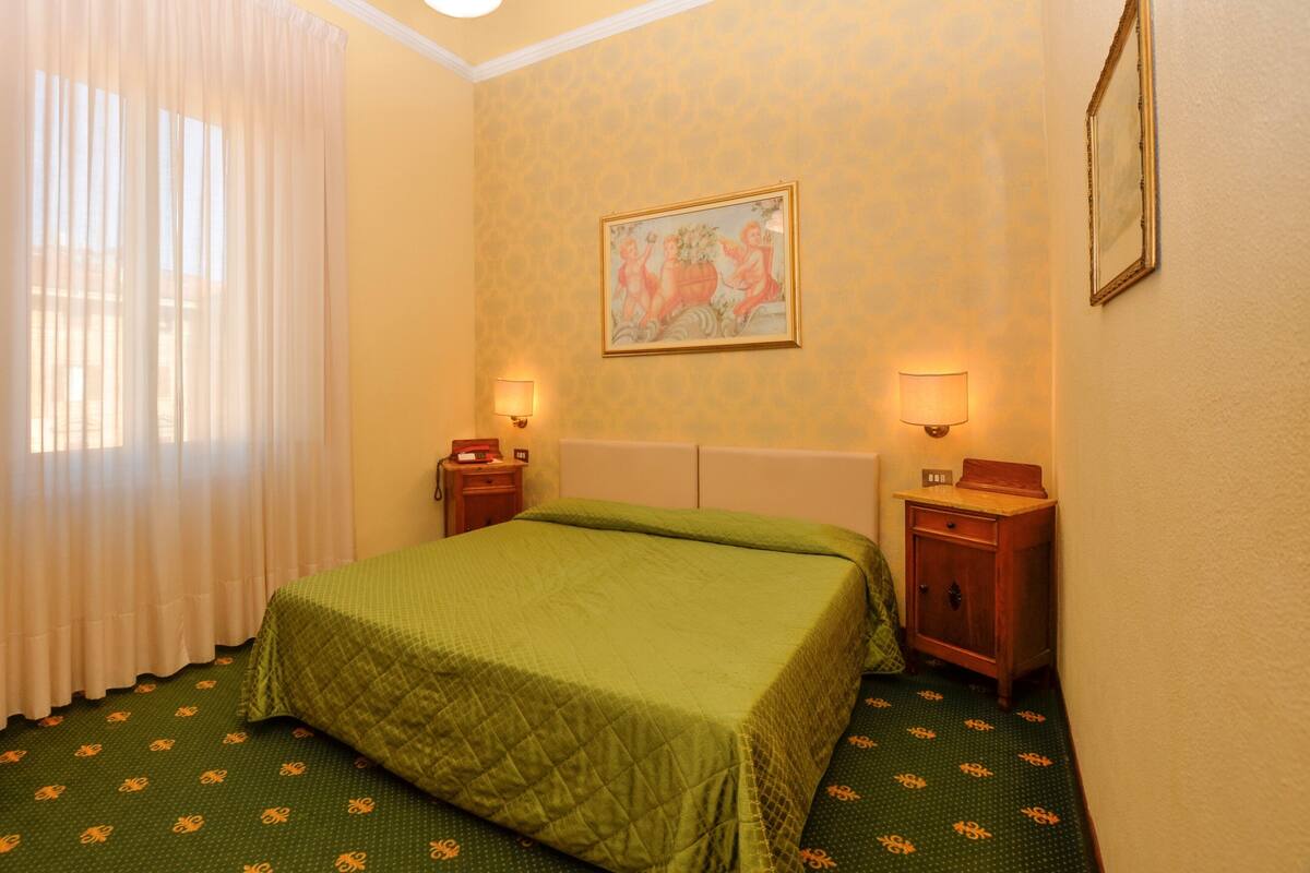 Habitación de hotel con cama, dos mesitas de noche, un cuadro en la pared y ventana con cortinas.