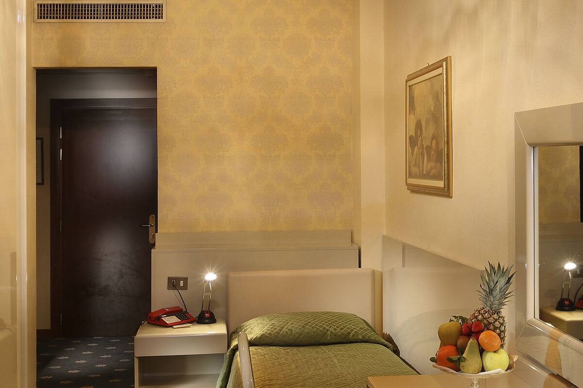 Una habitación de hotel con una cama, un escritorio, una silla, una pequeña mesa con fruta, un espejo y una araña.