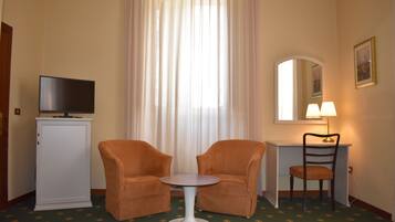 Une chambre d’hôtel avec deux fauteuils orange, une petite table blanche, un téléviseur posé sur un meuble, un bureau avec une lampe et un miroir.