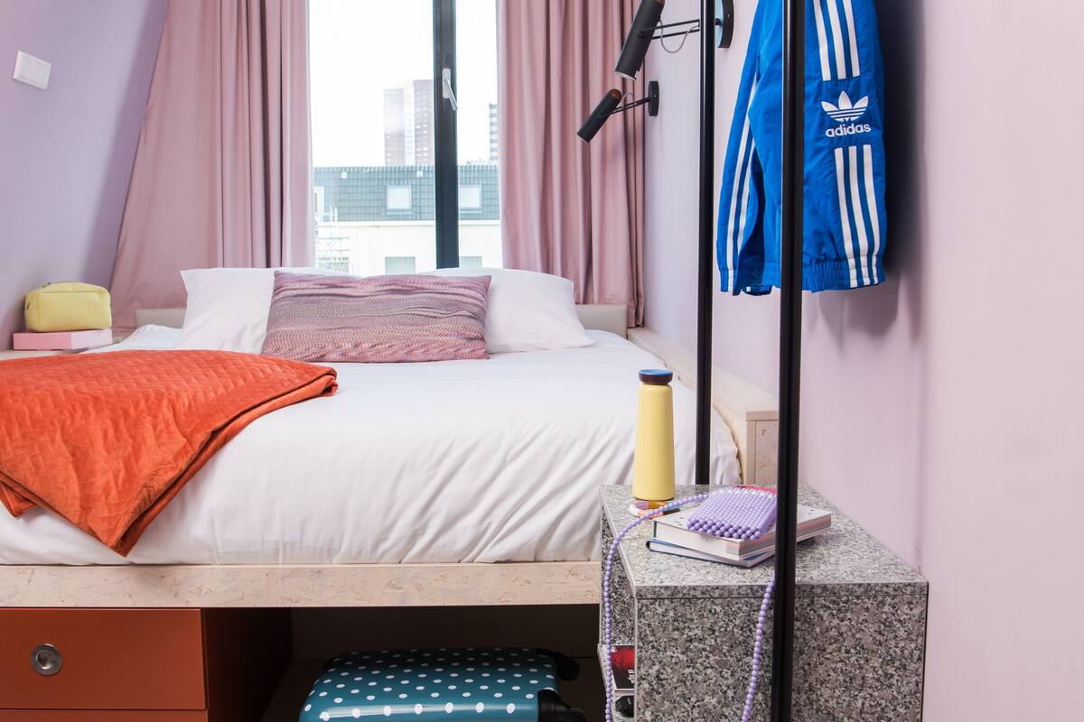 A bedroom with a bed, a suitcase, slippers, a jacket hanger, and a window with curtains.