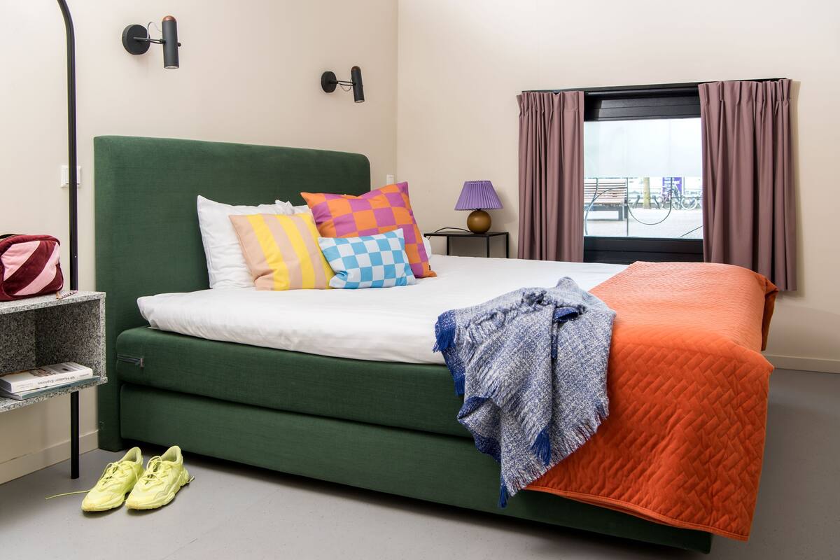 A neatly made bed with a green headboard, colorful pillows, and a blanket. There is a pair of yellow shoes on the floor, a small table with a lamp, and a window with curtains.
