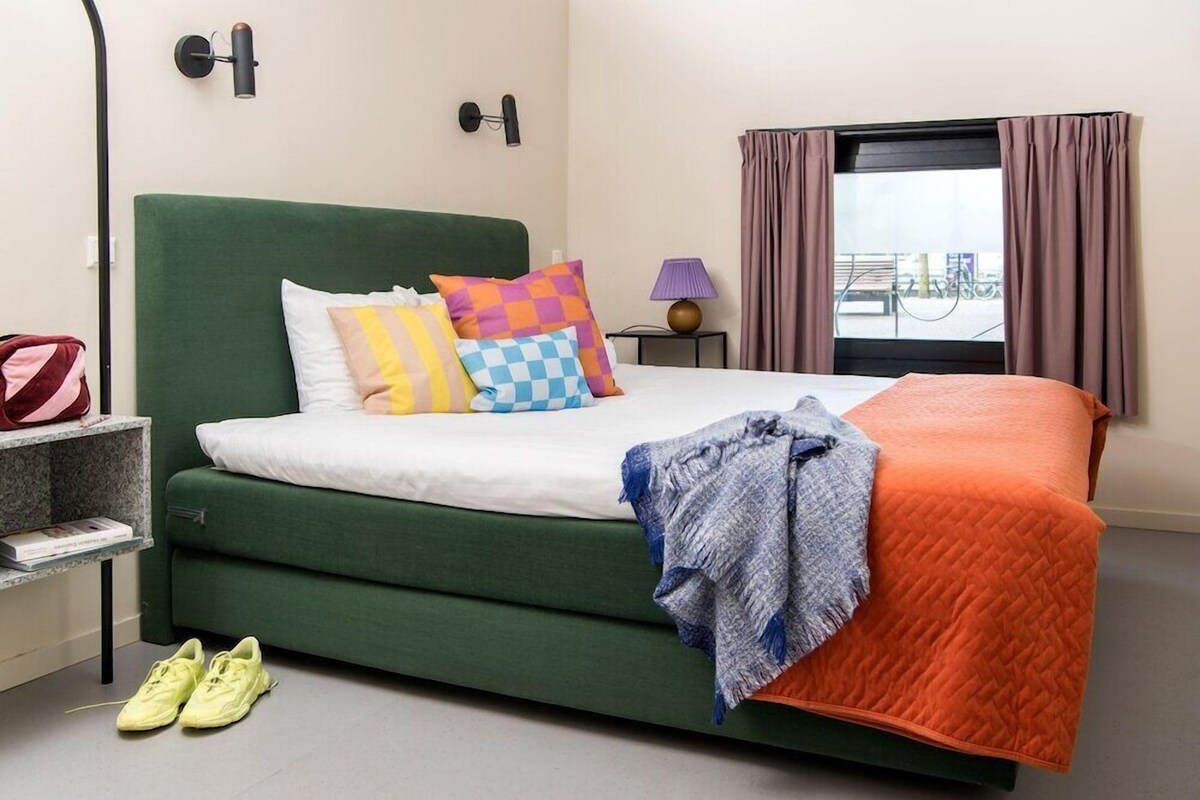 A neatly arranged hotel room with a green upholstered bed, colorful pillows, a grey blanket, and a view of the outdoors through a window with curtains.