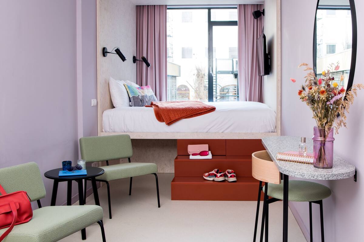 A modern hotel room with a bed, a desk, a chair, a small table, and a purple rug.