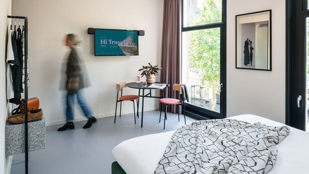 A hotel room with a bed, a desk with a chair, a window with curtains, a coat rack with clothes, and a TV displaying 'Hi Traveller,'. - hotel emma next (Rotterdam)