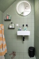 Un baño con un lavamanos blanco, un espejo redondo, un bote de basura negro y un perchero con una toalla a rayas.