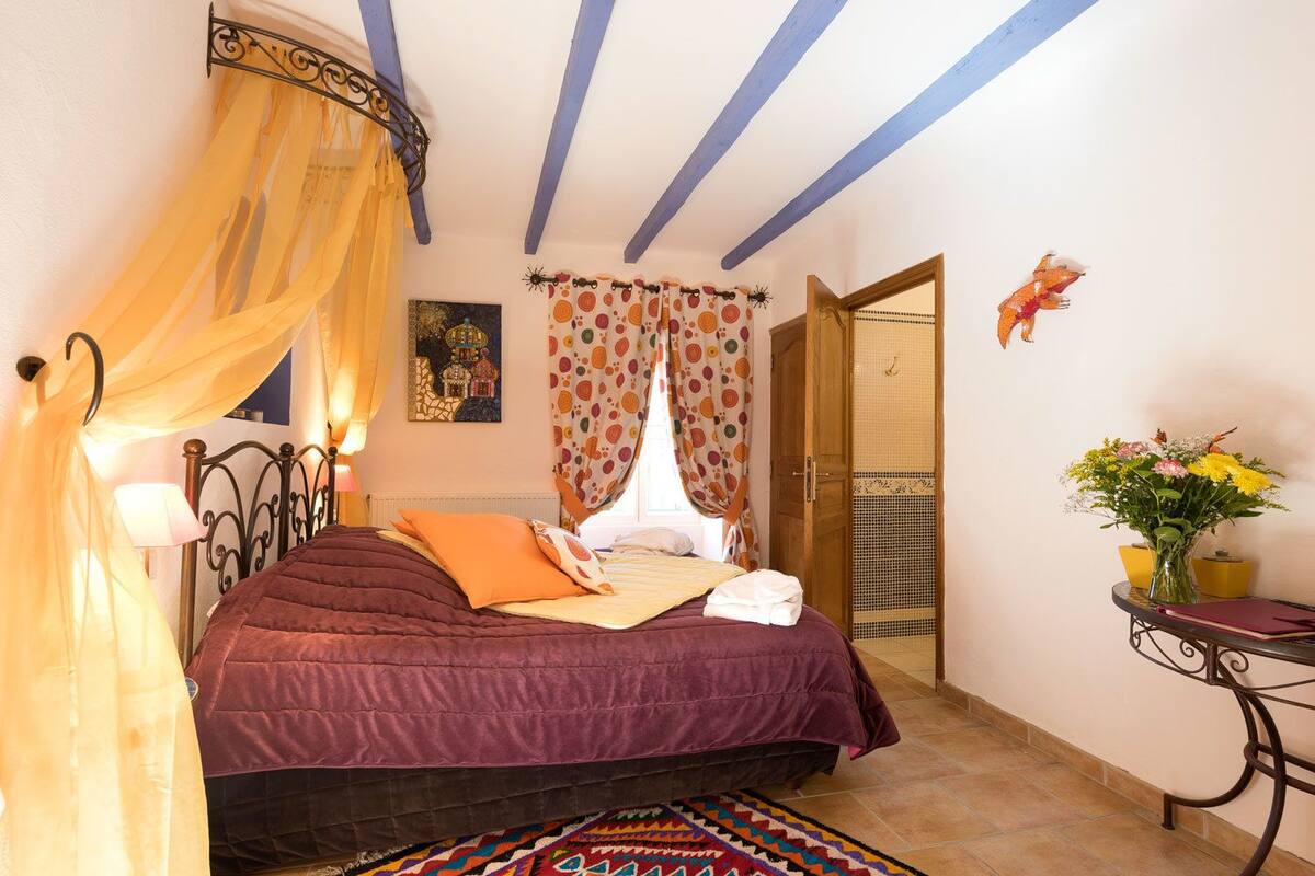 A bedroom with a bed, a nightstand, a bedside table, a window with curtains, a door, and a small table with a vase of flowers.