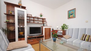 A modern living room with a sofa, a glass coffee table, a TV, wooden shelves, and a painting on the wall. - Apartment Ivanka (Kastela)