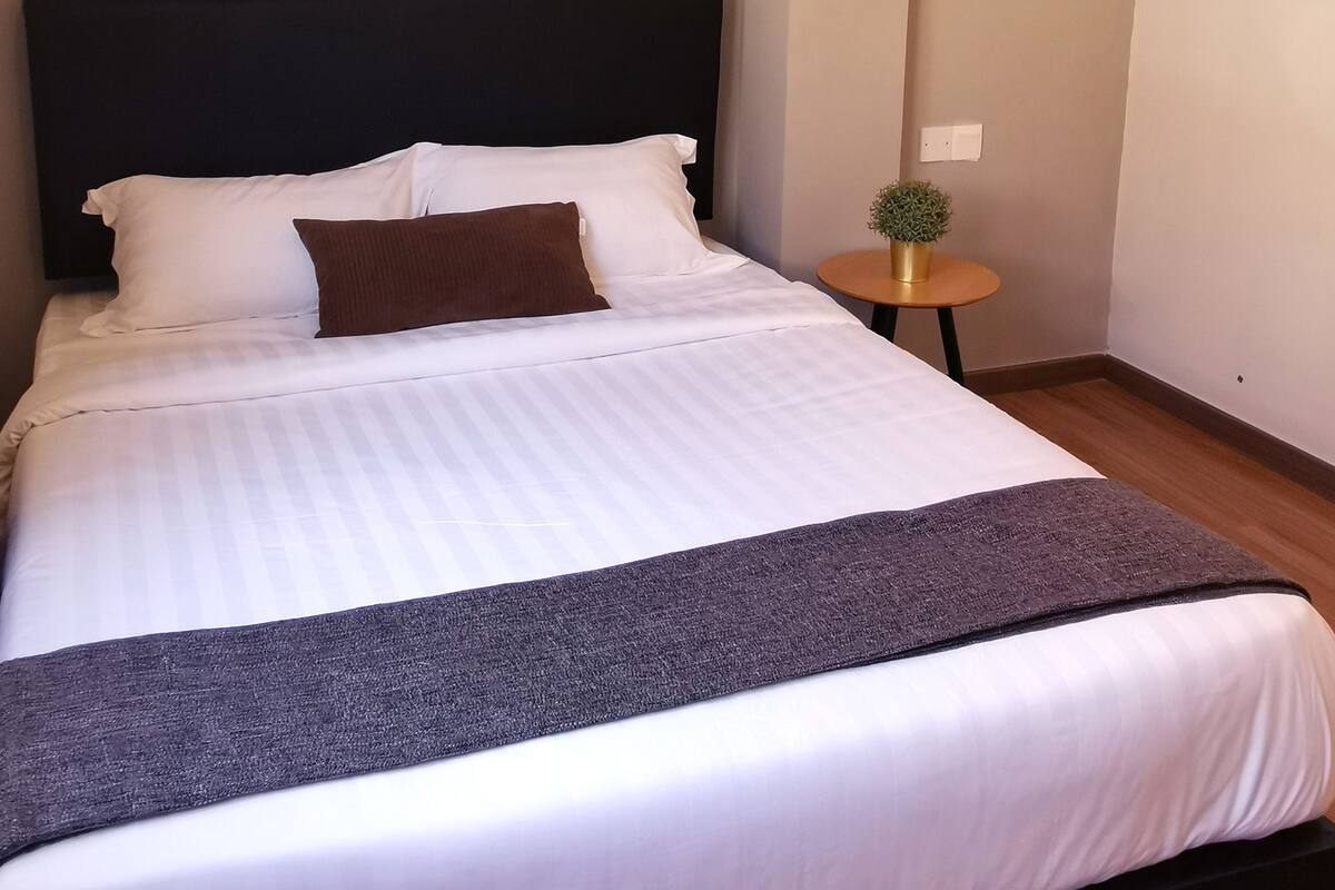 A neatly made bed with white and brown bedding, a dark headboard, and a small round table with a plant beside it.