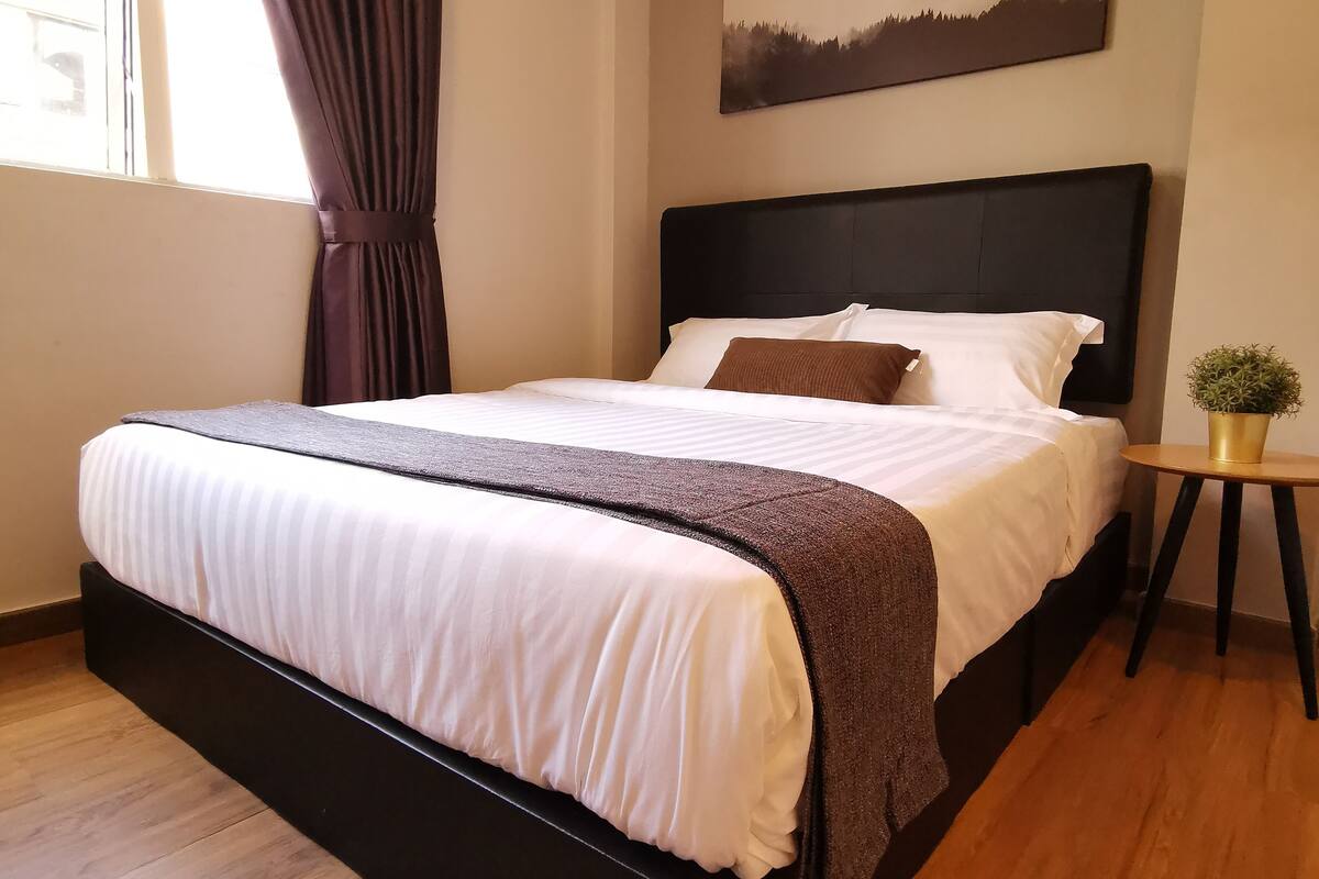 A modern bedroom with a large bed, a headboard, a small round table with a plant, and a painting on the wall.