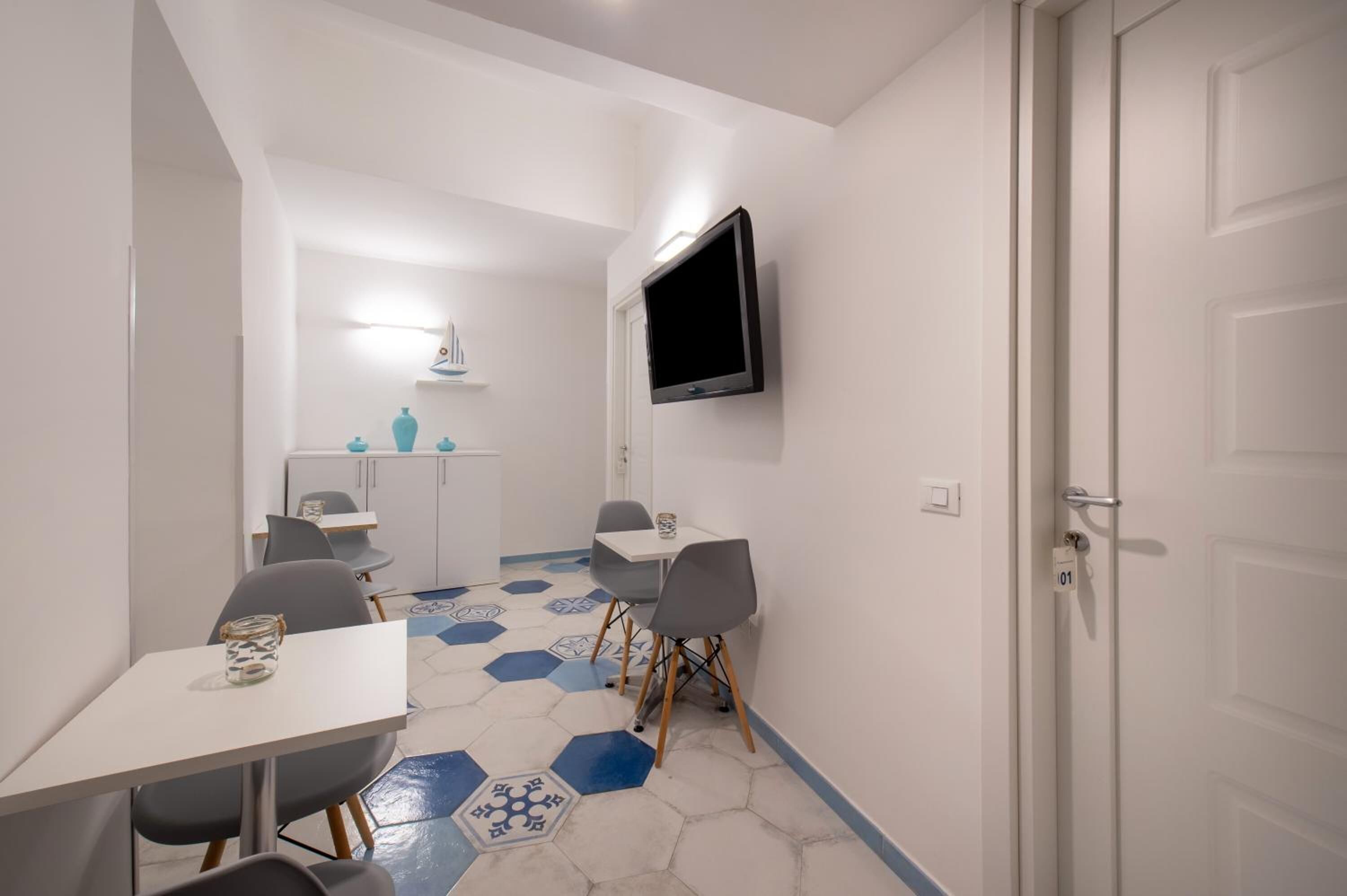 Habitación moderna y bien iluminada con decoración en tonos blancos, que incluye un área de comedor con piso de patrón hexagonal, un televisor de pantalla plana en la pared y una puerta con el rótulo '01'.