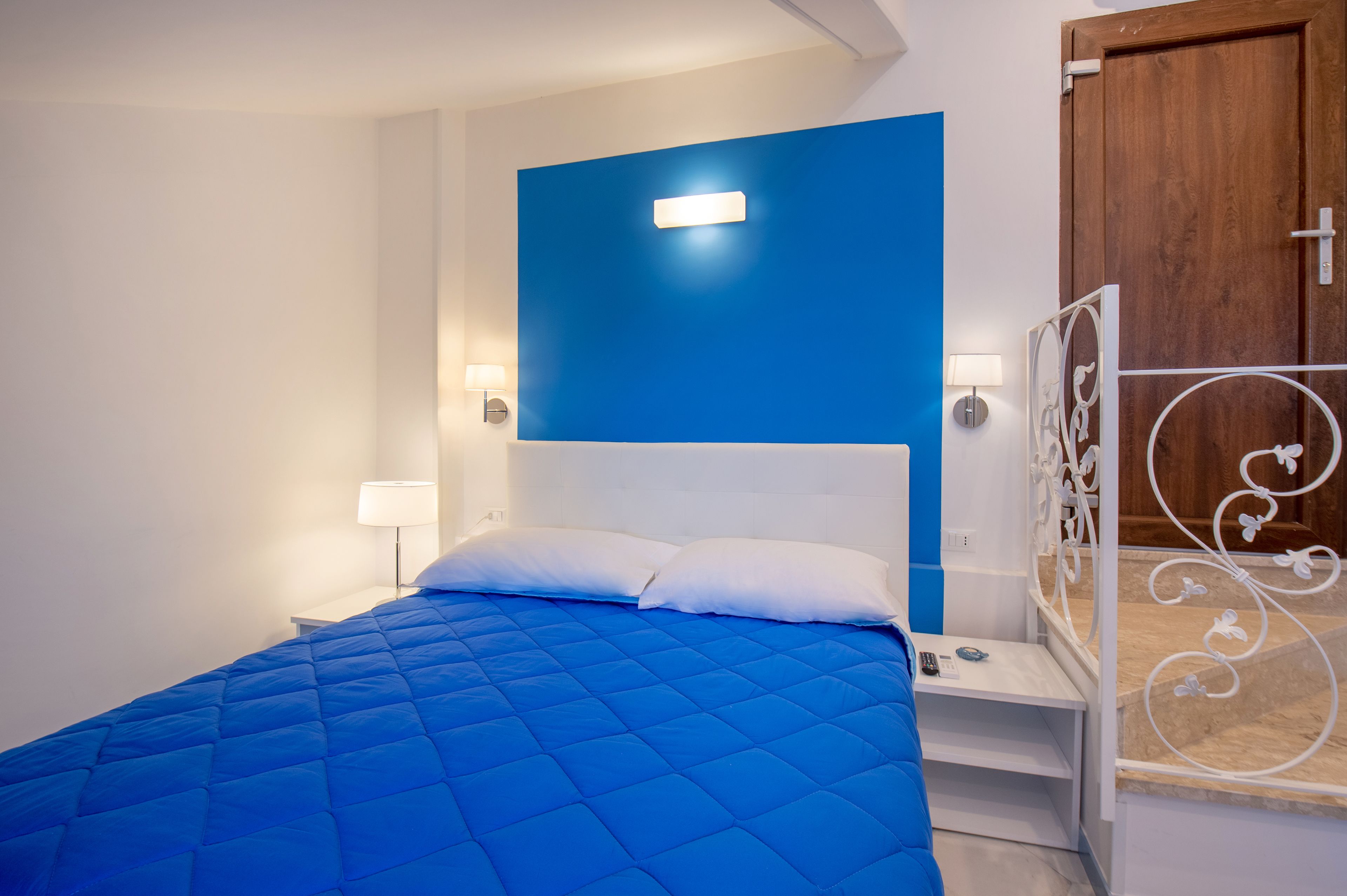 Un dormitorio con edredón azul acolchado, una mesita de noche blanca con lámpara y una puerta de madera con tirador de metal.