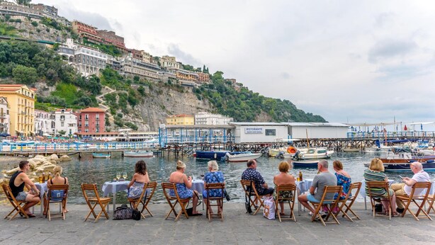 A waterfront dining area with tables and chairs, overlooking a marina with boats and a hillside of buildings. - Caruso Sea View (Sorrento)