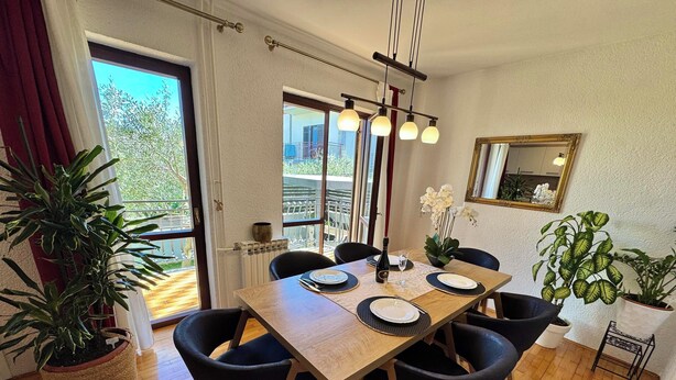 Un comedor con mesa de madera, sillas negras, plantas en macetas, una araña y vistas a través de la puerta de cristal.