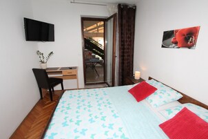 A bedroom with a bed, a desk, a chair, a TV mounted on the wall, and a door leading to a balcony.