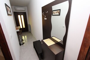 A room with a wooden vanity, a mirror, and a bench. There is a door with a picture frame and a light fixture above it.