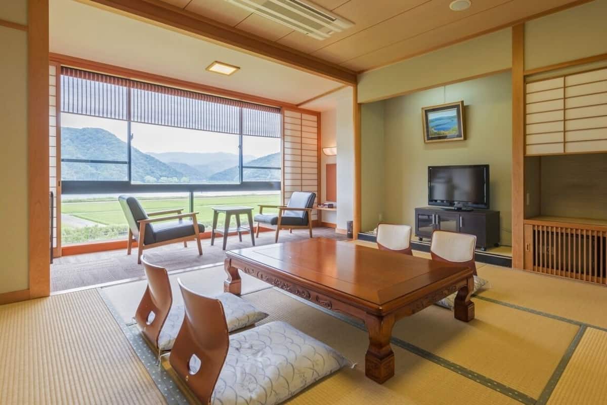 A traditional Japanese room with tatami flooring, a wooden table, chairs, a television, and a view of the outdoors.