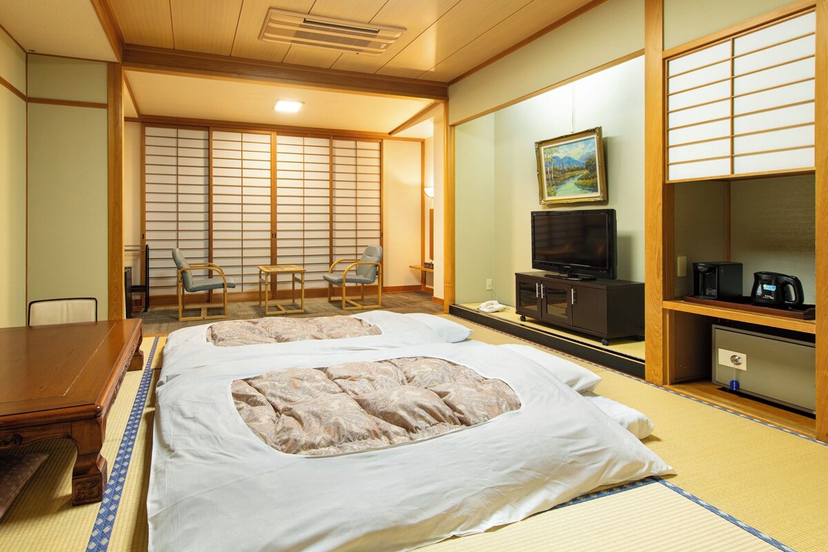 Ein traditionell eingerichtetes Hotelzimmer im japanischen Stil mit Tatami-Böden, einem Bett mit weißer und beiger Bettdecke, einem Fernseher, einer Kaffeemaschine und einem Bild an der Wand.