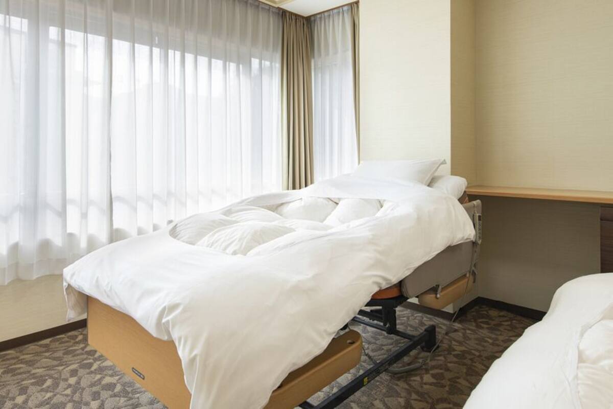 A hotel room with a bed, a wooden headboard, a window with sheer curtains, and a carpeted floor.