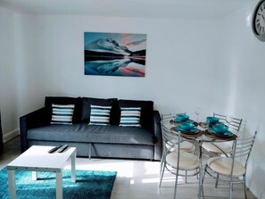 A modern living room with a grey sofa, a white coffee table, and a dining table set with blue bowls and glasses. - Walter Place (Edgware)